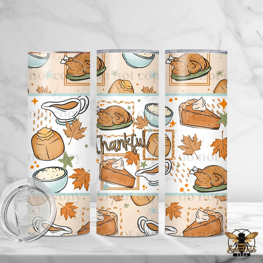 Three insulated tumblers with a Thanksgiving-themed pattern featuring roasted turkey, pie, pumpkin, maple leaves, gravy, and the word 'Thankful' on a light cream background. A clear lid is shown in the lower left on a white marble surface.