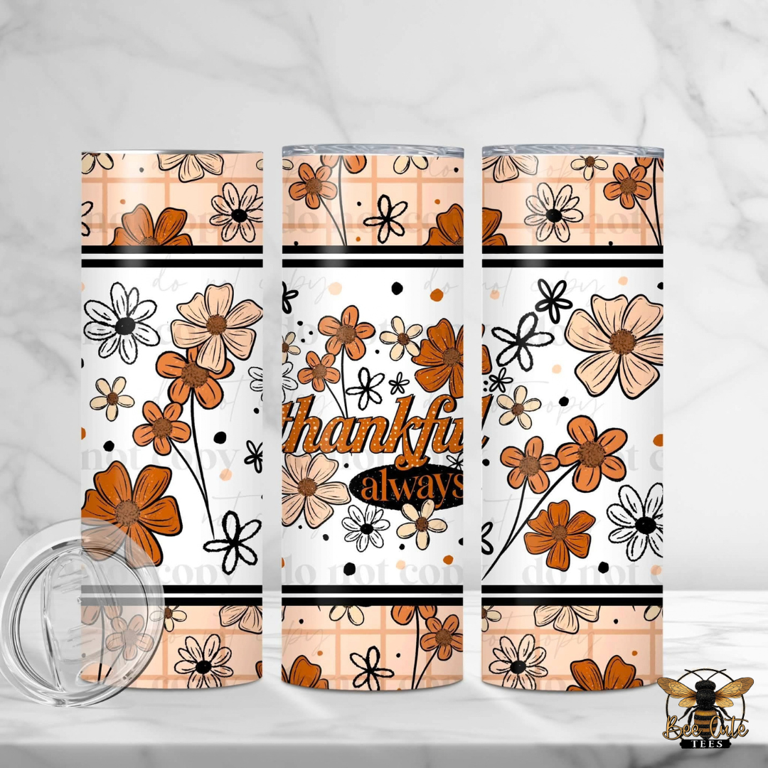 Three insulated tumblers with a floral pattern in orange, beige, and black on a white background, with one tumbler showing the text 'thankful always'. A clear lid is shown in the foreground on a white marble surface.