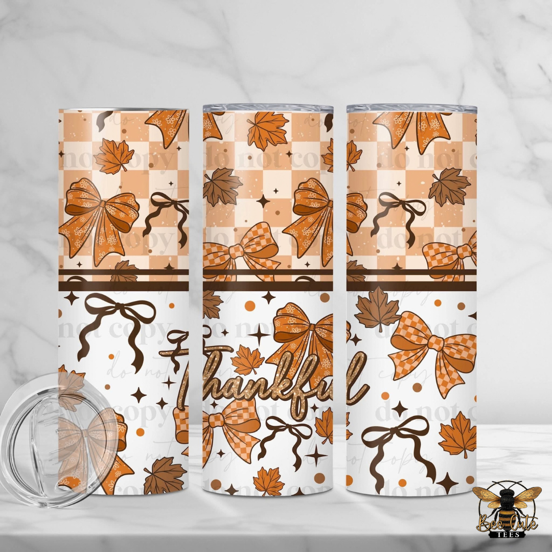 Three views of a skinny tumbler with a fall-themed pattern featuring orange and brown bows, maple leaves, stars, and the word 'Thankful' on a white background. A clear lid is shown in the bottom left corner on a white marble surface.