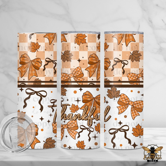 Three views of a skinny tumbler with a fall-themed pattern featuring orange and brown bows, maple leaves, stars, and the word 'Thankful' on a white background. A clear lid is shown in the bottom left corner on a white marble surface.