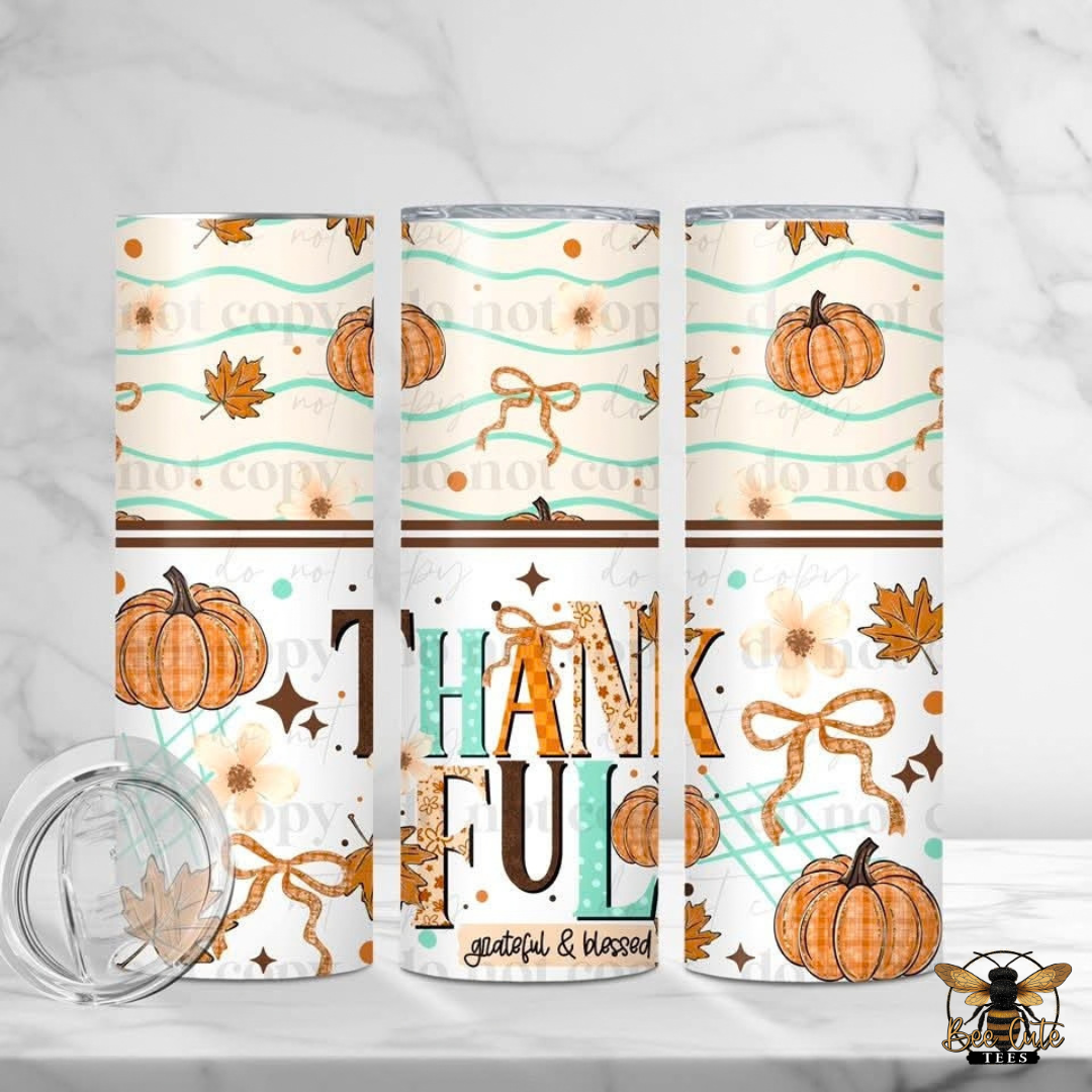 Three insulated tumblers with a fall-themed pattern featuring pumpkins, autumn leaves, bows, and the word 'THANKFUL' are displayed on a light marble surface with a clear lid in the foreground.