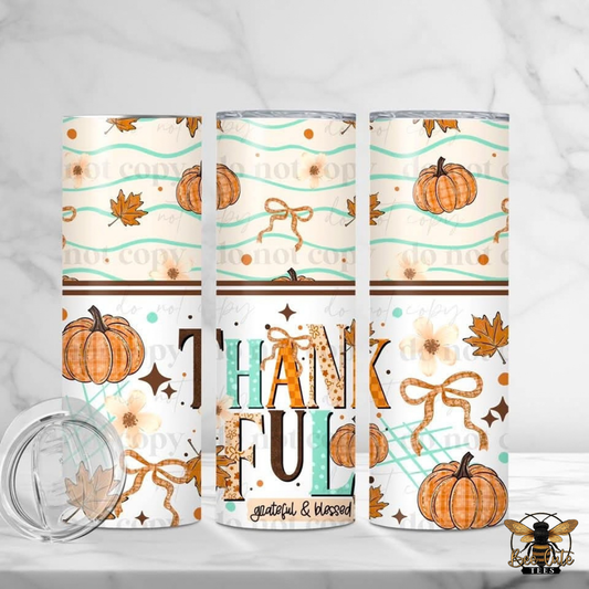 Three insulated tumblers with a fall-themed pattern featuring pumpkins, autumn leaves, bows, and the word 'THANKFUL' are displayed on a light marble surface with a clear lid in the foreground.