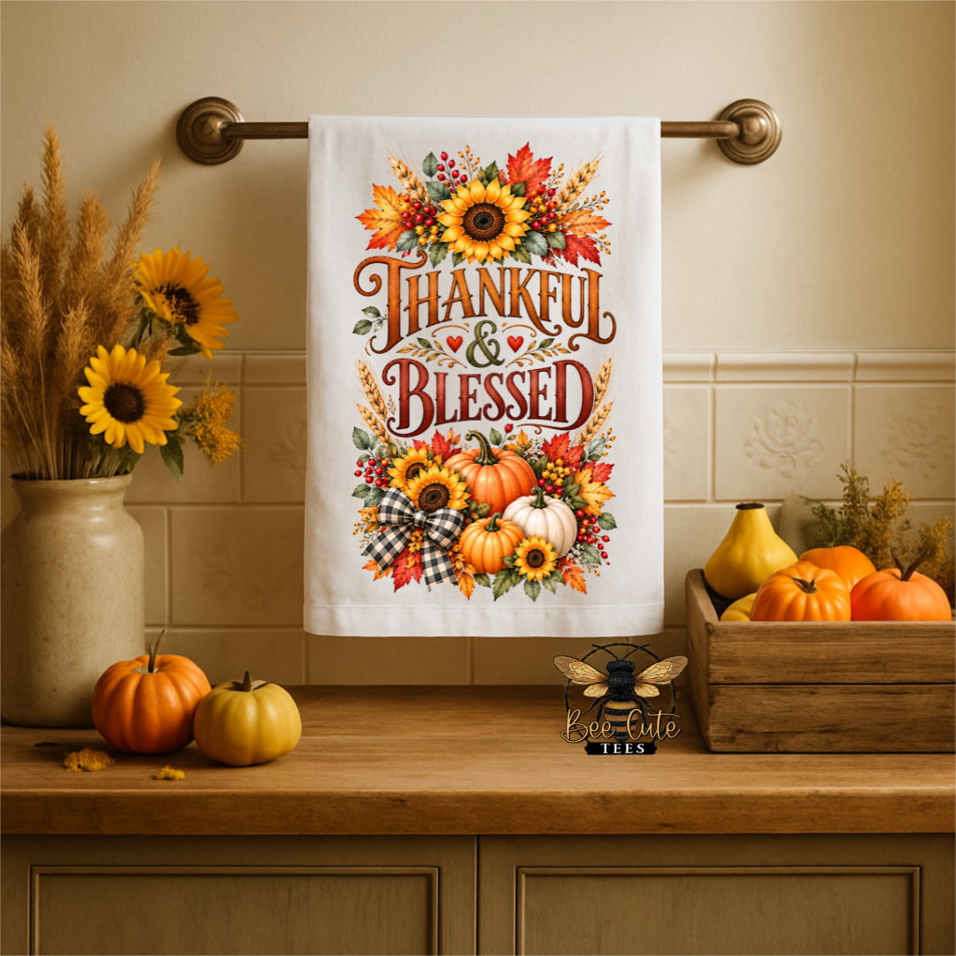 A white kitchen towel with a colorful autumn 'Thankful & Blessed' design hangs on a brass towel bar above a wooden counter decorated with pumpkins, sunflowers, and a wooden crate.