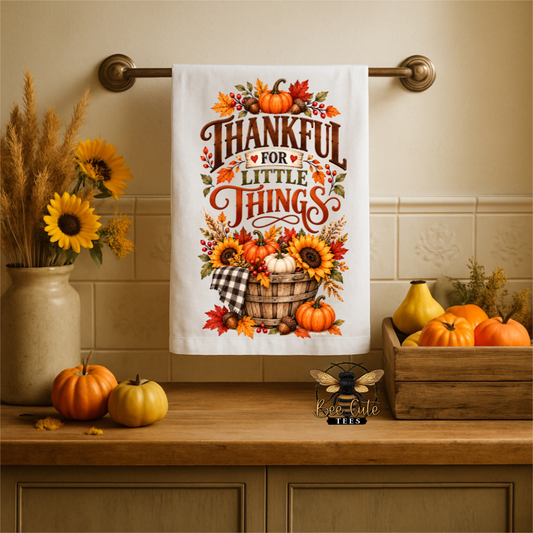A white kitchen towel with a colorful autumn 'Thankful for Little Things' print hangs on a brass towel bar over a wooden countertop. The scene is styled with fall decorations including pumpkins, sunflowers, and a wooden crate.