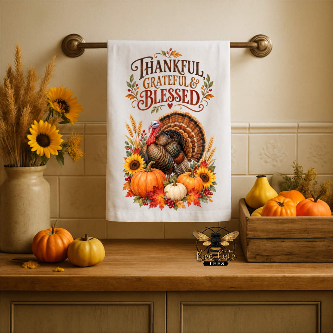 A white kitchen towel with a Thanksgiving design hangs on a brass towel bar. The towel features the text 'THANKFUL, GRATEFUL & BLESSED' above an illustration of a turkey surrounded by pumpkins, sunflowers, and autumn leaves.