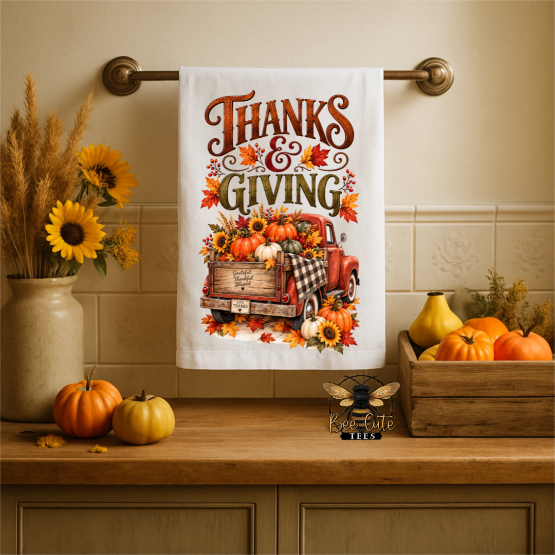 A white kitchen towel with a 'Thanks & Giving' autumn graphic hangs on a brass towel bar. The towel features a red pickup truck carrying pumpkins and sunflowers, surrounded by orange and yellow leaves.