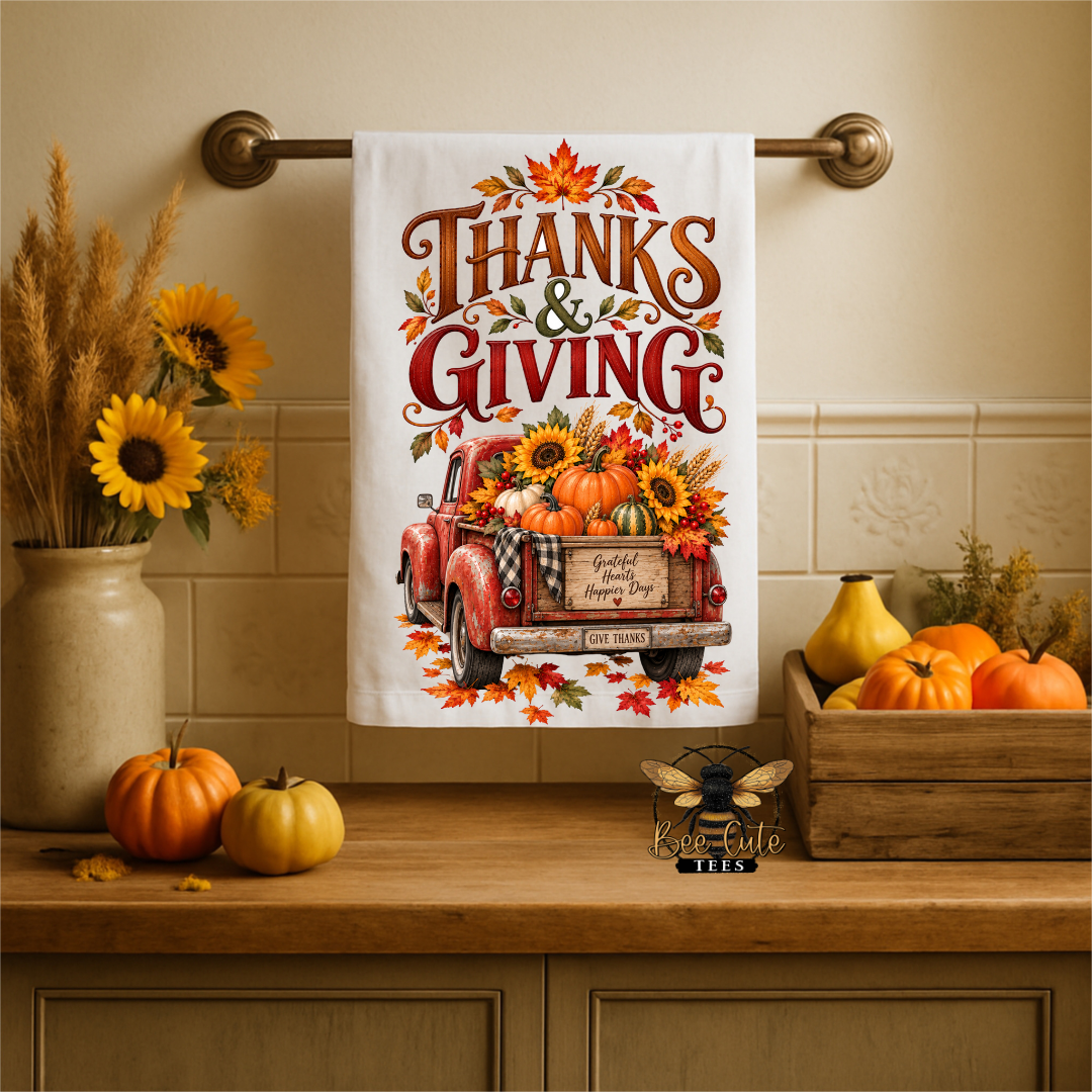 A white kitchen towel with a Thanksgiving-themed autumn print hangs on a brass towel bar. The towel features the words 'THANKS & GIVING' above an illustration of a red pickup truck loaded with pumpkins and sunflowers, with falling leaves around it.