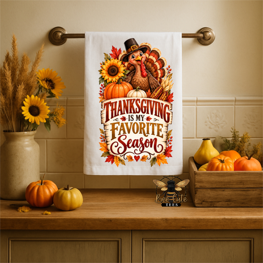 A white kitchen towel hanging on a brass rack above a wooden counter, printed with a Thanksgiving-themed turkey, sunflowers, pumpkins, and the phrase 'Thanksgiving is my favorite season.' The scene is styled with fall decor, including sunflowers, pumpkins, and a wooden crate, and the brand name 'bee Cute Tees' appears near the bottom.