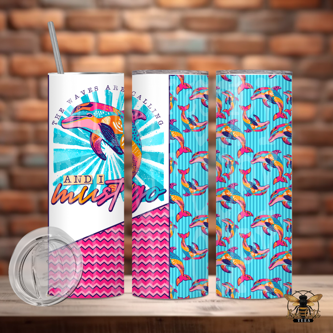 Colorful 20 oz tumbler featuring bright dolphin artwork, tropical patterns, and beach-inspired text that reads “The Waves Are Calling And I Must Go,” displayed with a lid and metal straw against a rustic brick background.
