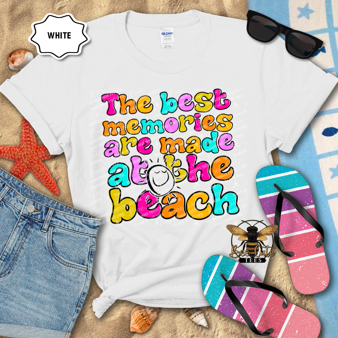White t-shirt with colorful beach-themed text, sunglasses, and flip-flops on a sandy background.