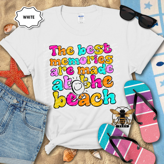 White t-shirt with colorful beach-themed text, sunglasses, and flip-flops on a sandy background.