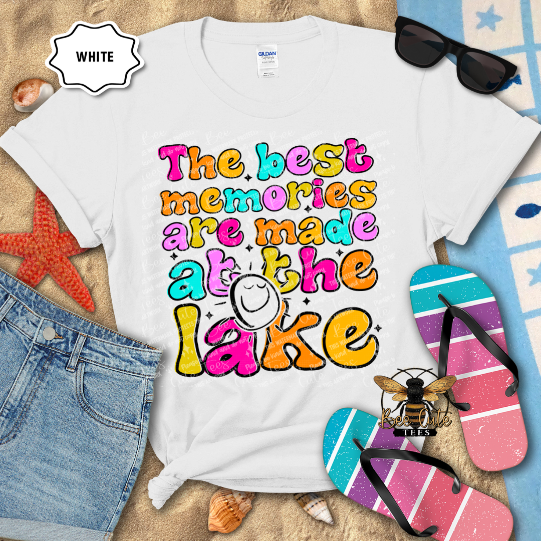 White t-shirt with colorful text 'The best memories are made at the lake' on a beach-themed background.