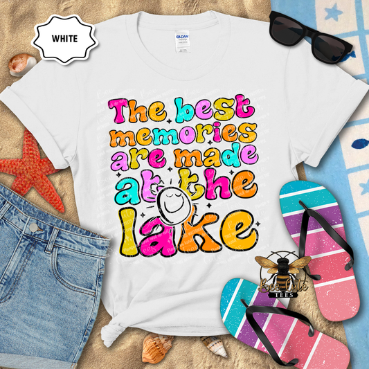 White t-shirt with colorful text 'The best memories are made at the lake' on a beach-themed background.