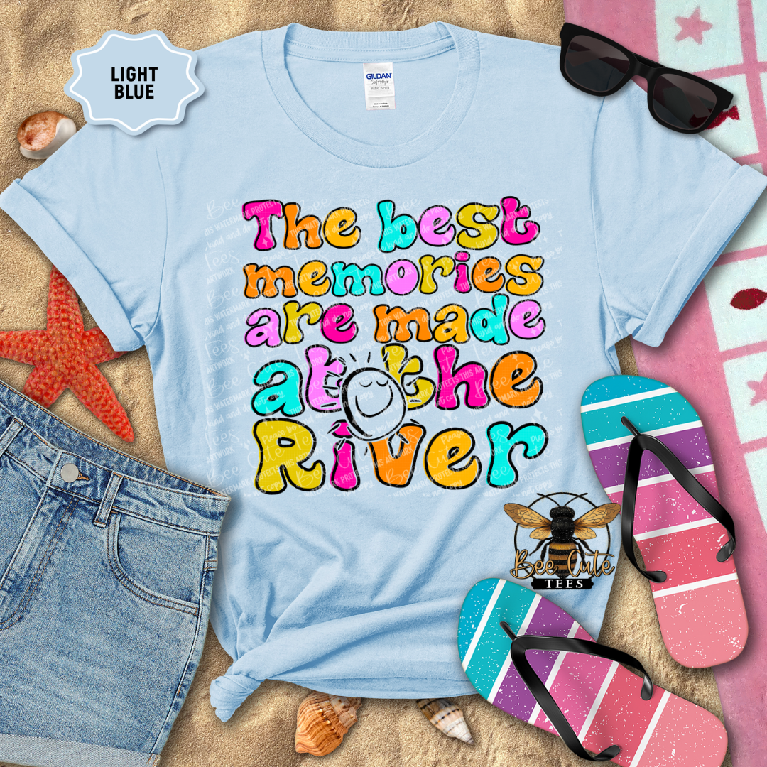 Light blue t-shirt with colorful text 'The best memories are made at the River' on a beach-themed background.