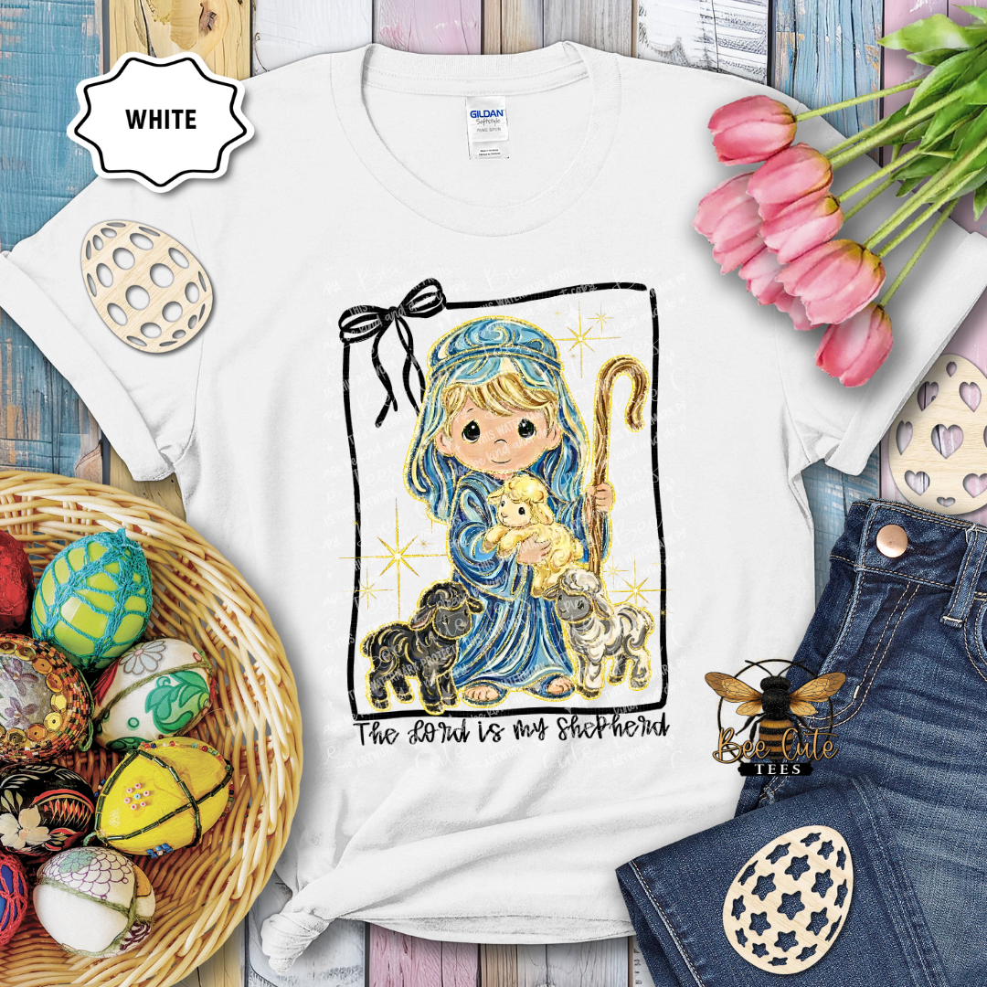 White t-shirt with a religious design featuring a shepherd and lamb, surrounded by Easter decorations.