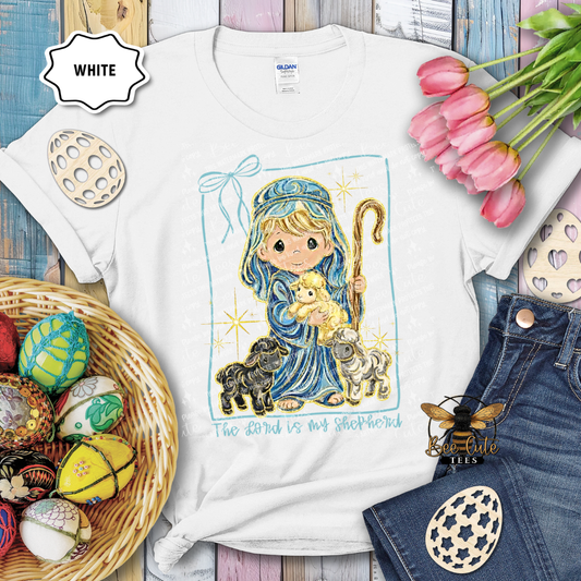 White t-shirt with a child holding a lamb design, surrounded by Easter decorations on a wooden surface.