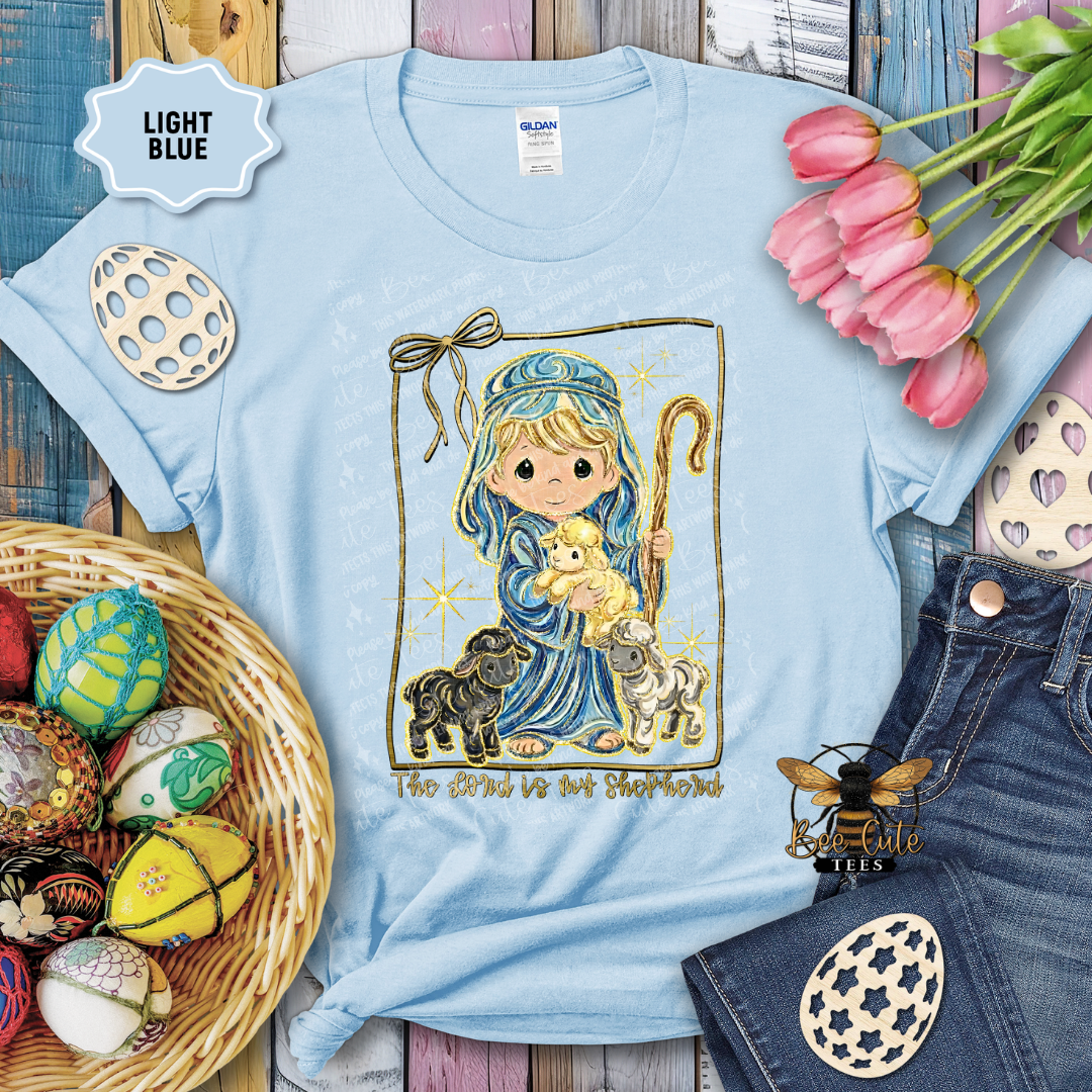 Light blue t-shirt with a cartoon design of a shepherd holding a teddy bear, surrounded by Easter decorations.