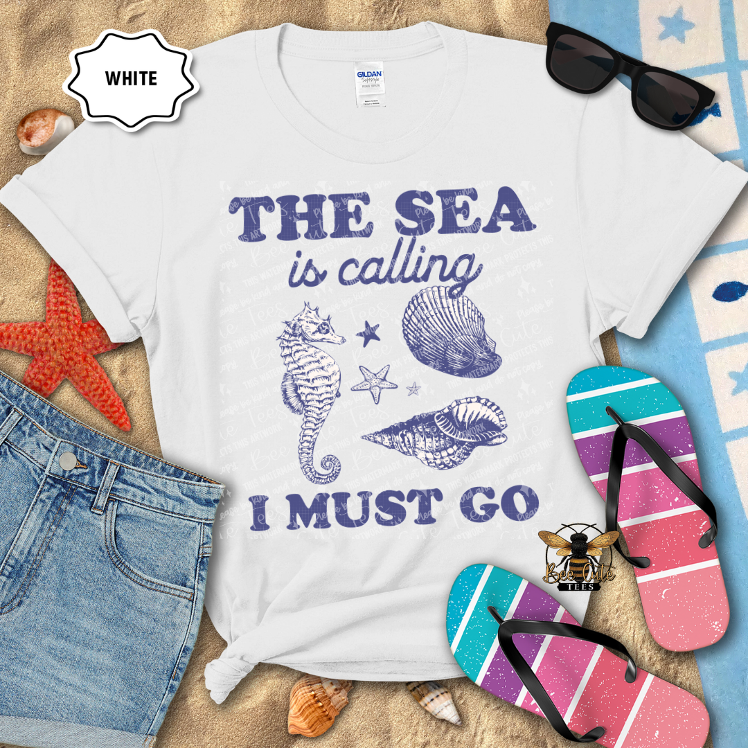 White t-shirt with 'The Sea is Calling I Must Go' text and beach-themed graphics on a sandy background with flip-flops and sunglasses.