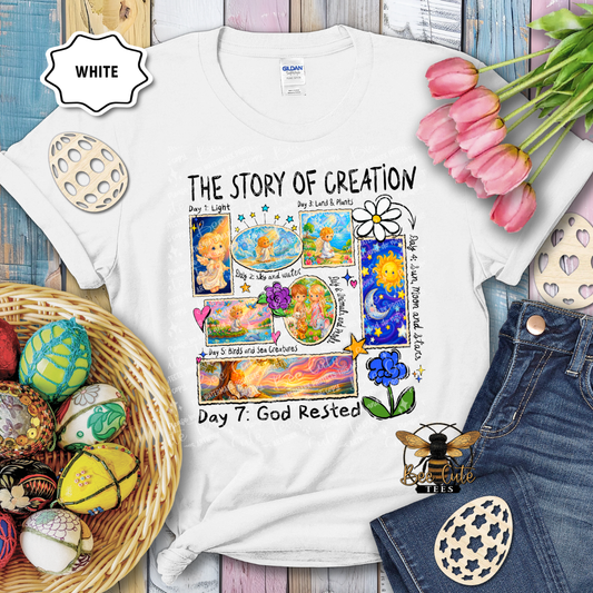 White t-shirt with 'The Story of Creation' design, surrounded by Easter eggs and flowers on a wooden surface.
