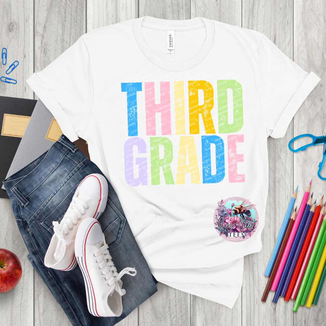 White t-shirt with 'THIRD GRADE' in colorful letters, jeans, sneakers, and school supplies on a wooden surface.