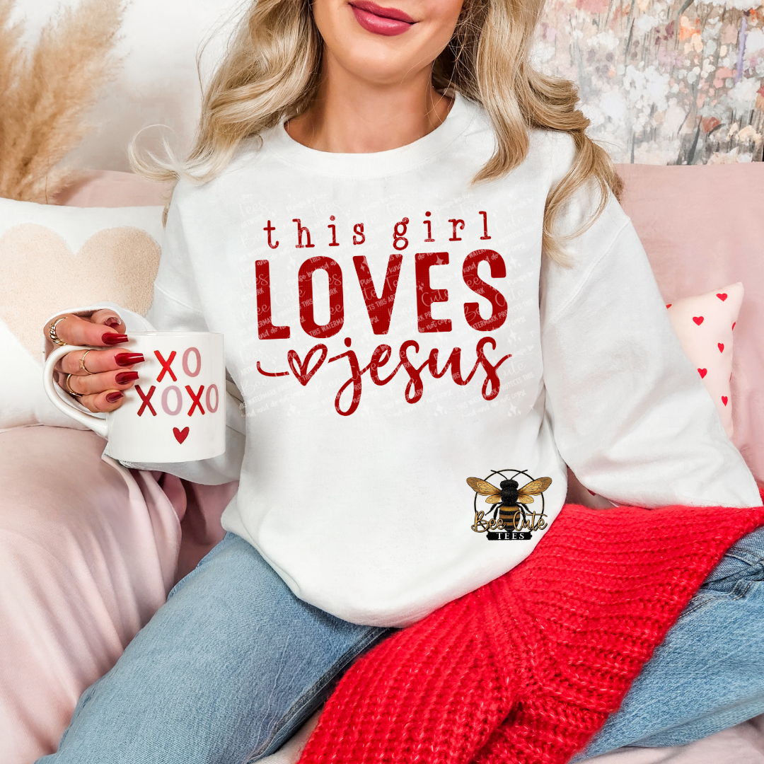 White crewneck sweatshirt featuring red “this girl LOVES Jesus” graphic with heart detail, styled on a model holding a mug in a cozy, feminine setting with soft decor and a knit blanket.
