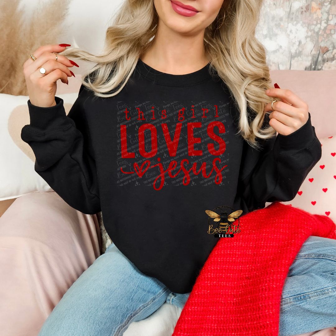 Black crewneck sweatshirt featuring red “this girl LOVES Jesus” graphic with heart detail, styled on a model holding a mug in a cozy, feminine setting with soft decor and a knit blanket.
