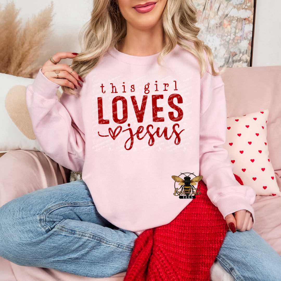 Pink crewneck sweatshirt featuring red “this girl LOVES Jesus” graphic with heart detail, styled on a model holding a mug in a cozy, feminine setting with soft decor and a knit blanket.
