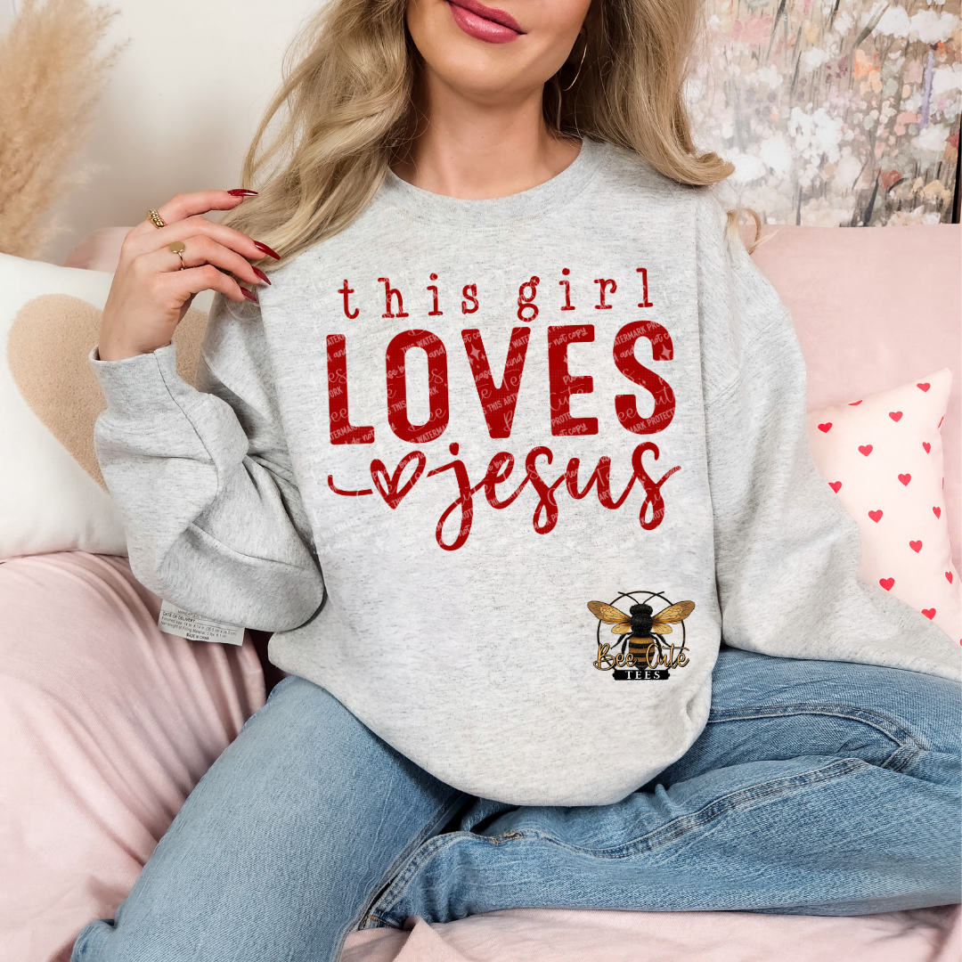 Grey crewneck sweatshirt featuring red “this girl LOVES Jesus” graphic with heart detail, styled on a model holding a mug in a cozy, feminine setting with soft decor and a knit blanket.
