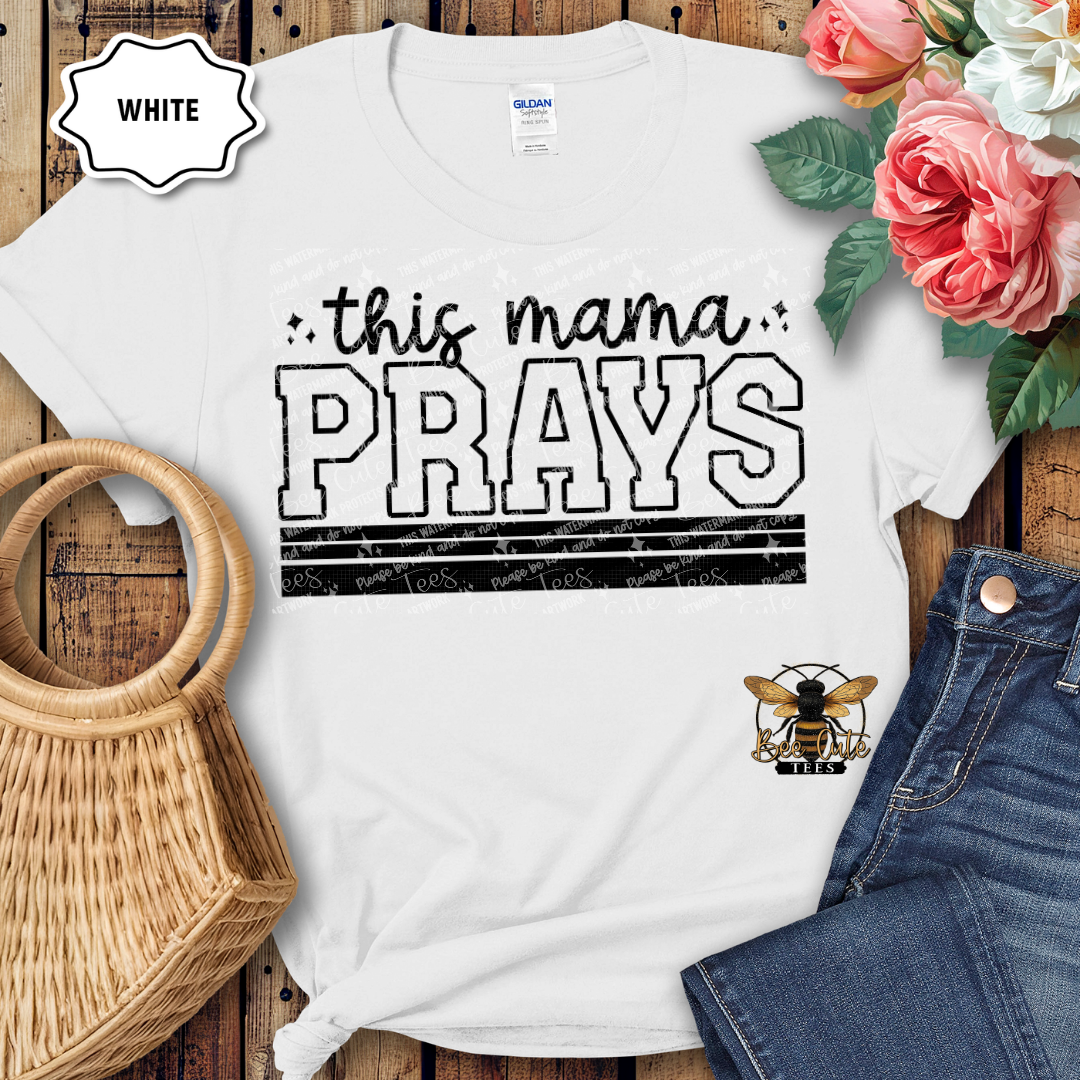 White t-shirt with 'this mama PRAYS' text on a wooden surface with flowers and jeans.