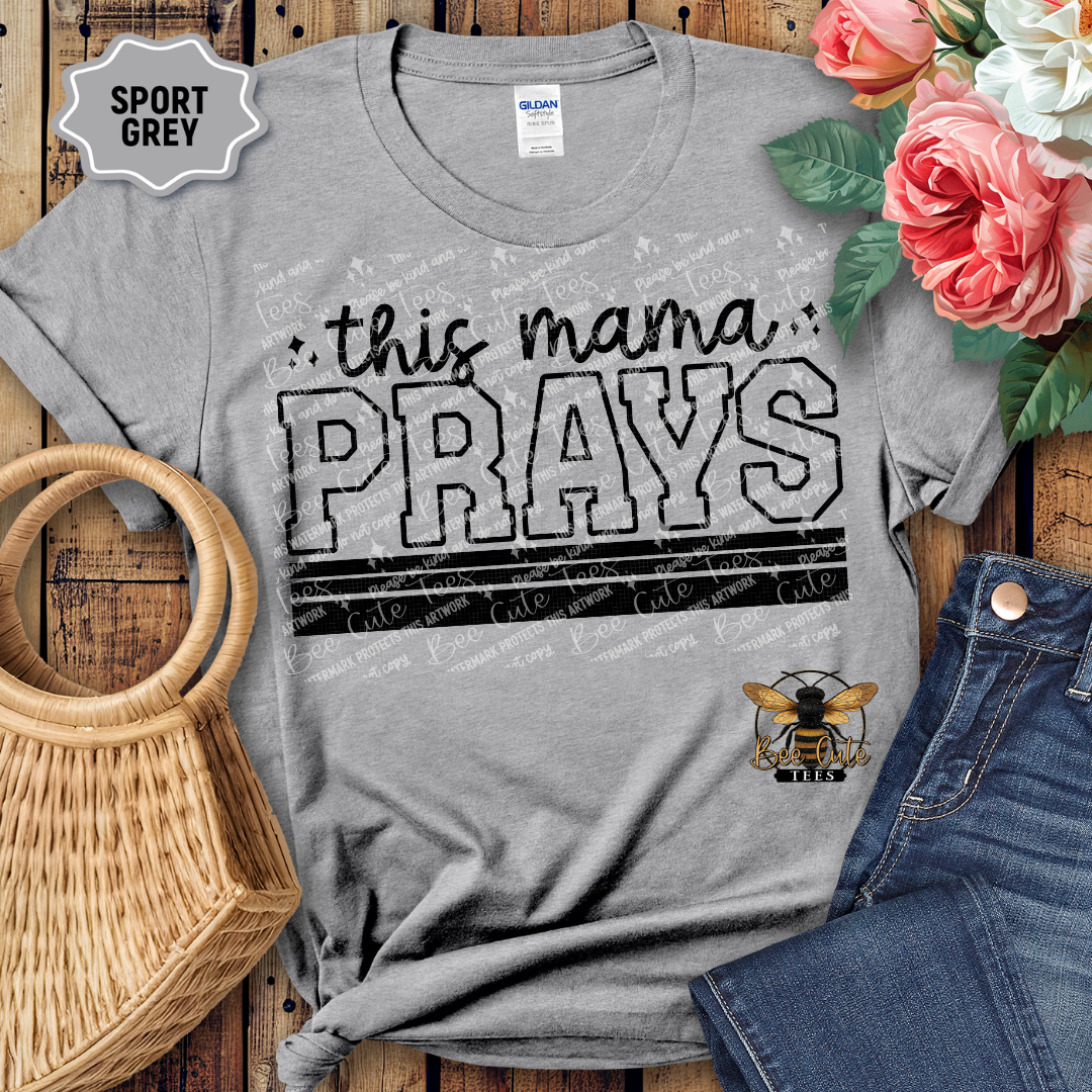 Gray t-shirt with 'this mama PRAYS' text and bee logo on a wooden surface with flowers and jeans.