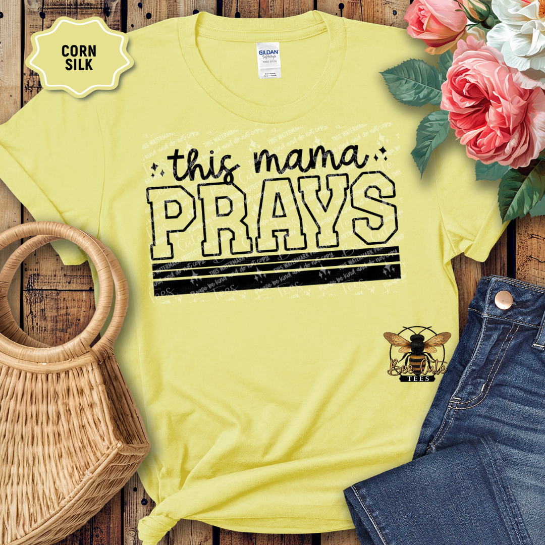 Yellow t-shirt with 'this mama PRAYS' text and a bee logo on a wooden surface with flowers and jeans.