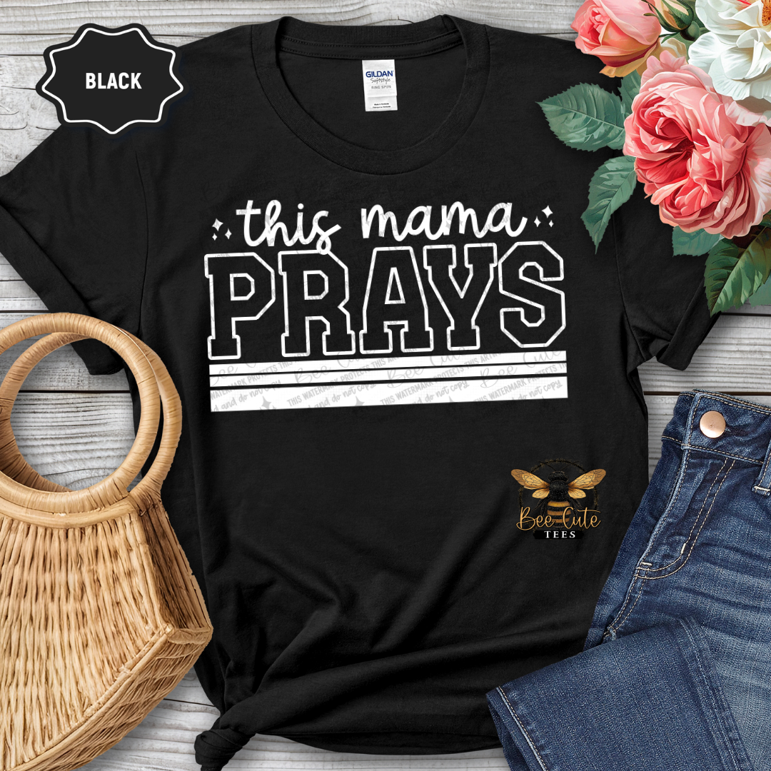 Black t-shirt with 'this mama PRAYS' text, surrounded by flowers and jeans on a wooden surface.