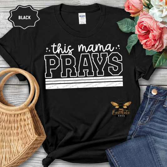 Black t-shirt with 'this mama PRAYS' text, surrounded by flowers and jeans on a wooden surface.