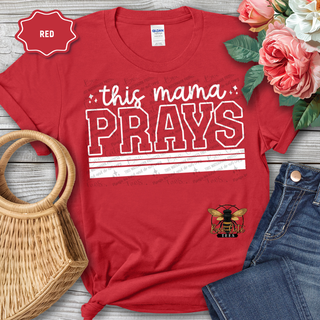 Red t-shirt with 'this mama PRAYS' text and floral and denim accessories on a wooden surface