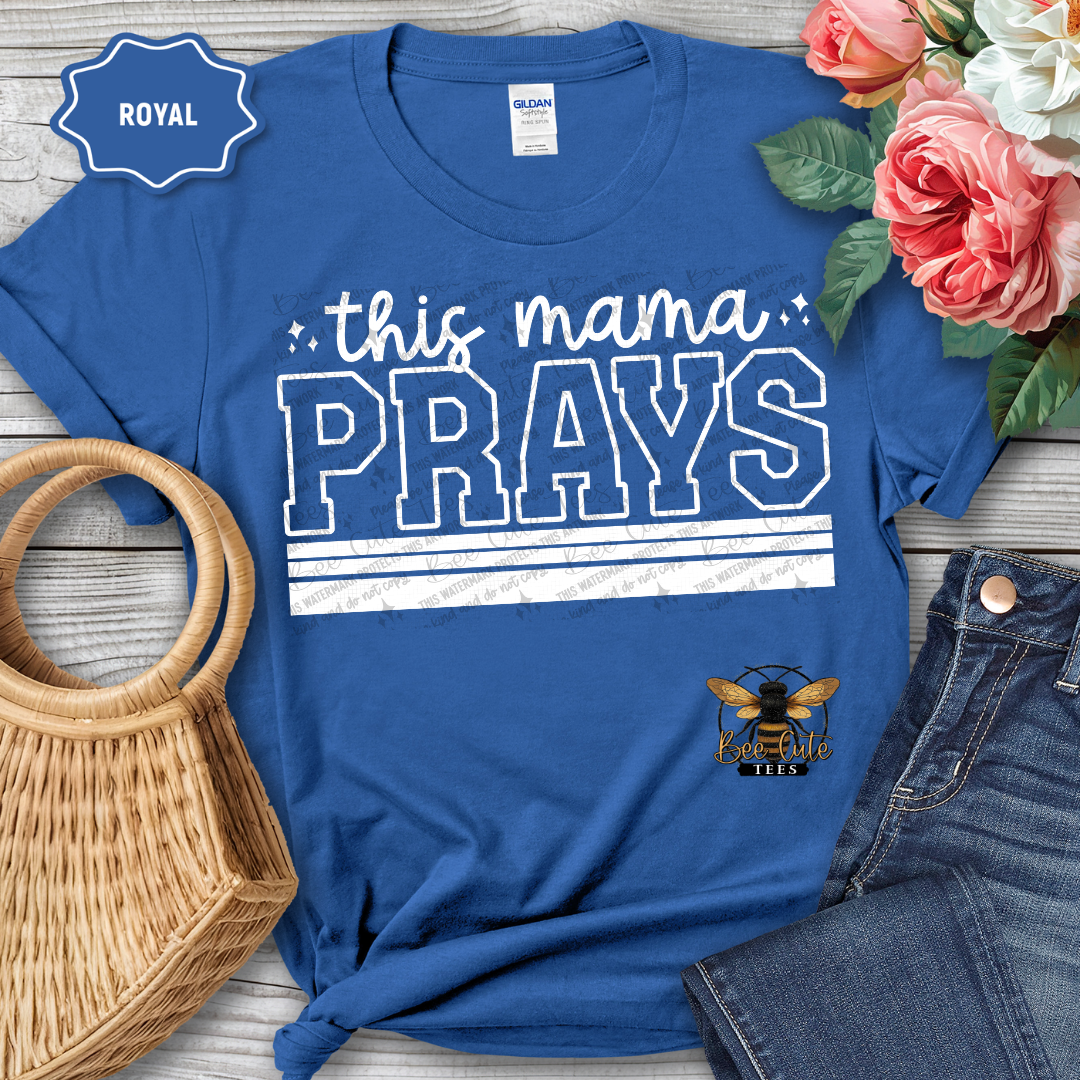 Blue t-shirt with 'this mama PRAYS' text and bee logo, placed on a wooden surface with flowers and accessories.