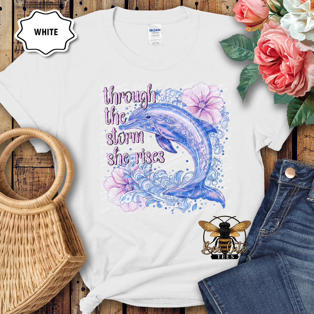 White t-shirt with a dolphin and floral design, text 'through the storm she rises', placed on a wooden surface with flowers and jeans.