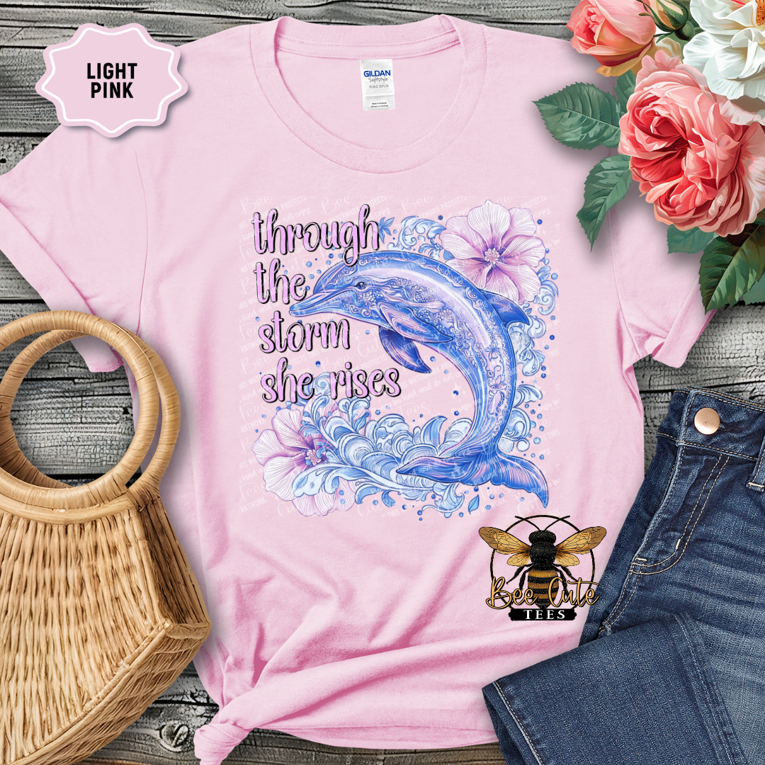 Pink t-shirt with dolphin design and text 'through the storm she rises' on a wooden surface with flowers and jeans.