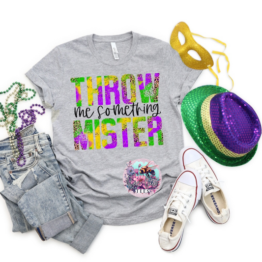 A gray t-shirt with the text 'THROW ME SOMETHING' printed on it, displayed alongside Mardi Gras-themed accessories including a hat, shoes, and beads.