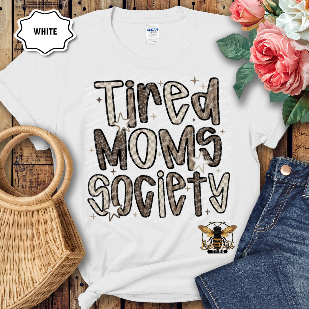 White t-shirt with 'Tired Moms Society' text and a bee logo on a wooden surface with flowers and jeans.