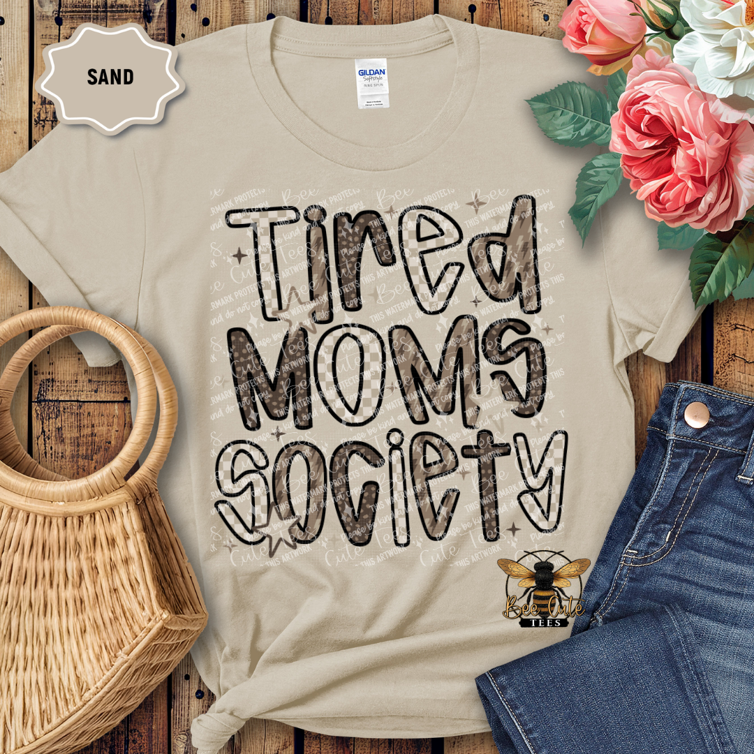 Beige t-shirt with 'Tired Moms Society' text on a wooden surface with flowers and jeans.