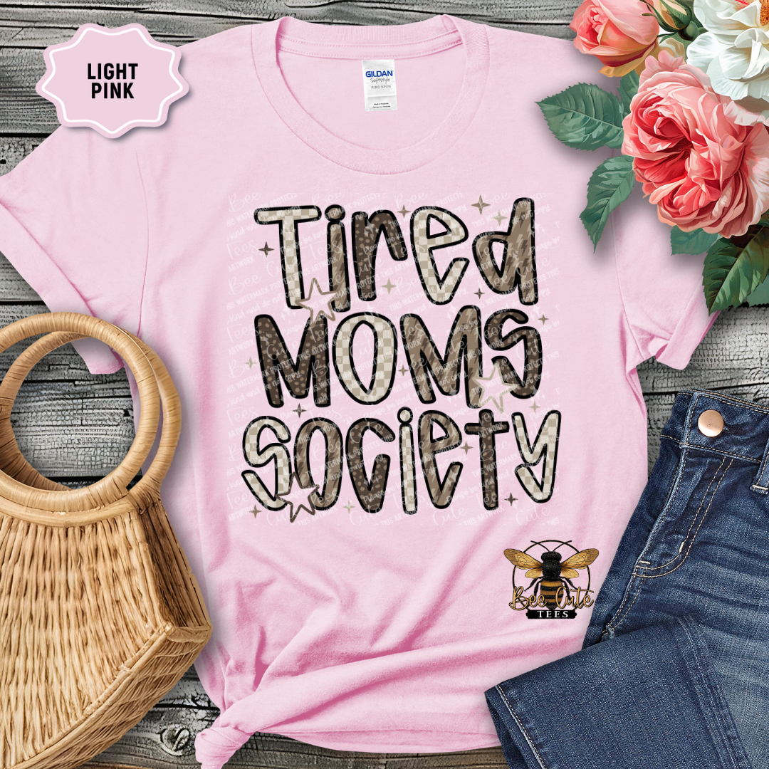 Light pink t-shirt with 'Tired Moms Society' text and a bee logo, placed on a wooden surface with flowers and denim jeans.