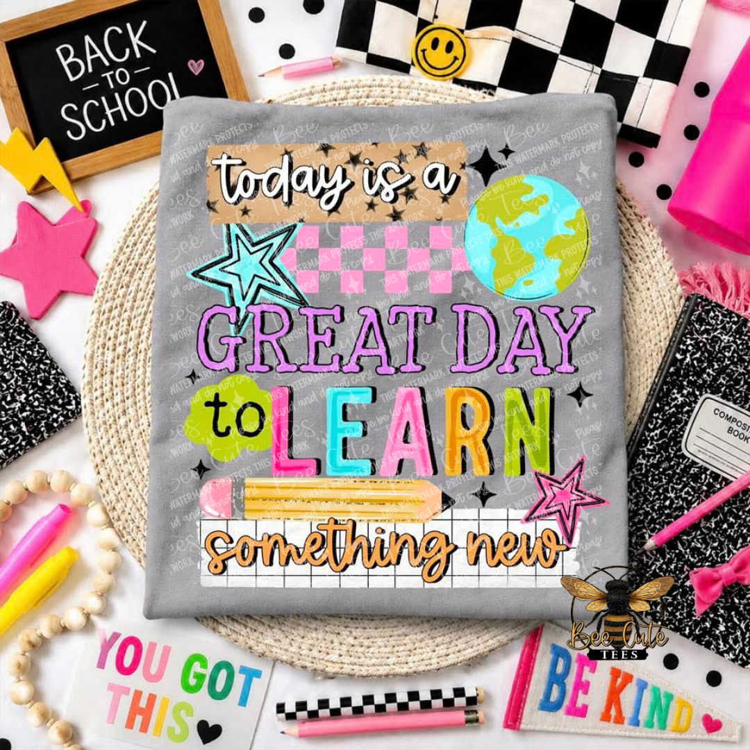 A folded  gray t-shirt, featuring a colorful school-themed graphic that reads 'today is a GREAT DAY to LEARN something new' with stars, a globe, and a pencil. The fabric is styled in a flat-lay arrangement with school supplies, notebooks, and a chalkboard.