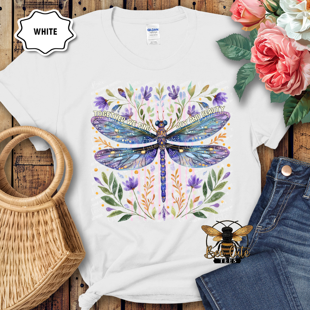 White t-shirt with a colorful dragonfly design on a wooden surface with flowers and jeans.