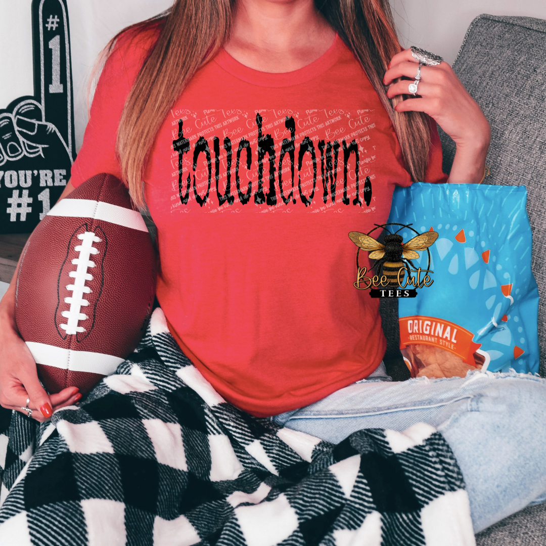 Red graphic t-shirt featuring distressed black “touchdown.” text, shown on a model holding a football and snack bag, styled for a casual game day look.
