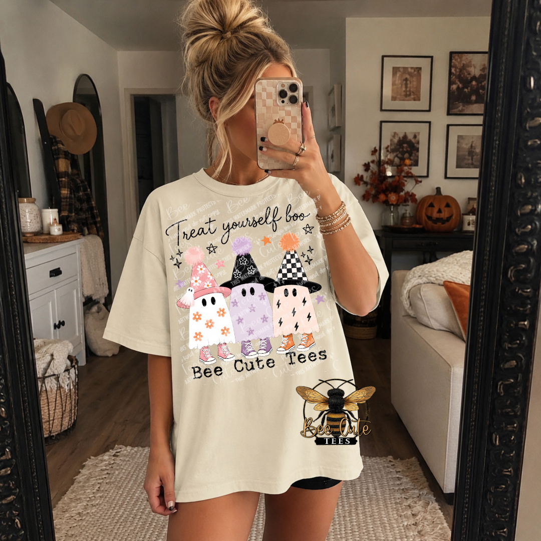 Woman taking a mirror selfie wearing an oversized cream short-sleeve tee featuring three adorable pastel ghosts in pink, lavender, and peach, each wearing a colorful witch hat and matching sneakers. Black script reads “Treat Yourself Boo” above the ghosts, with “Bee Cute Tees” printed below. Styled in a cozy bedroom decorated for fall.