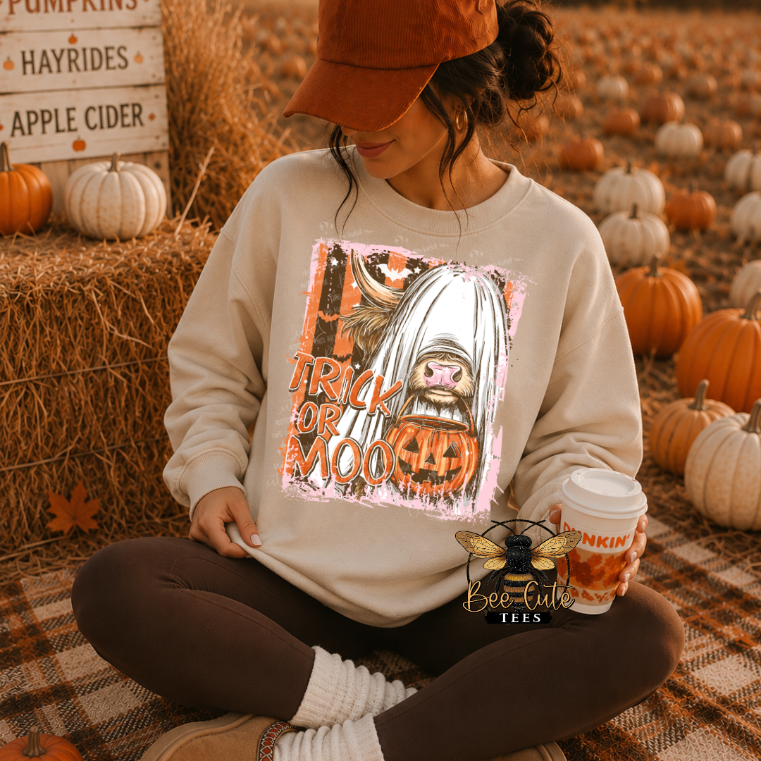 A woman sits cross-legged on a plaid blanket in a pumpkin patch setting, wearing a cream sweatshirt with a 'Trick or Treat' cartoon graphic, a brown cap, and brown leggings. She holds a pumpkin-themed coffee cup.
