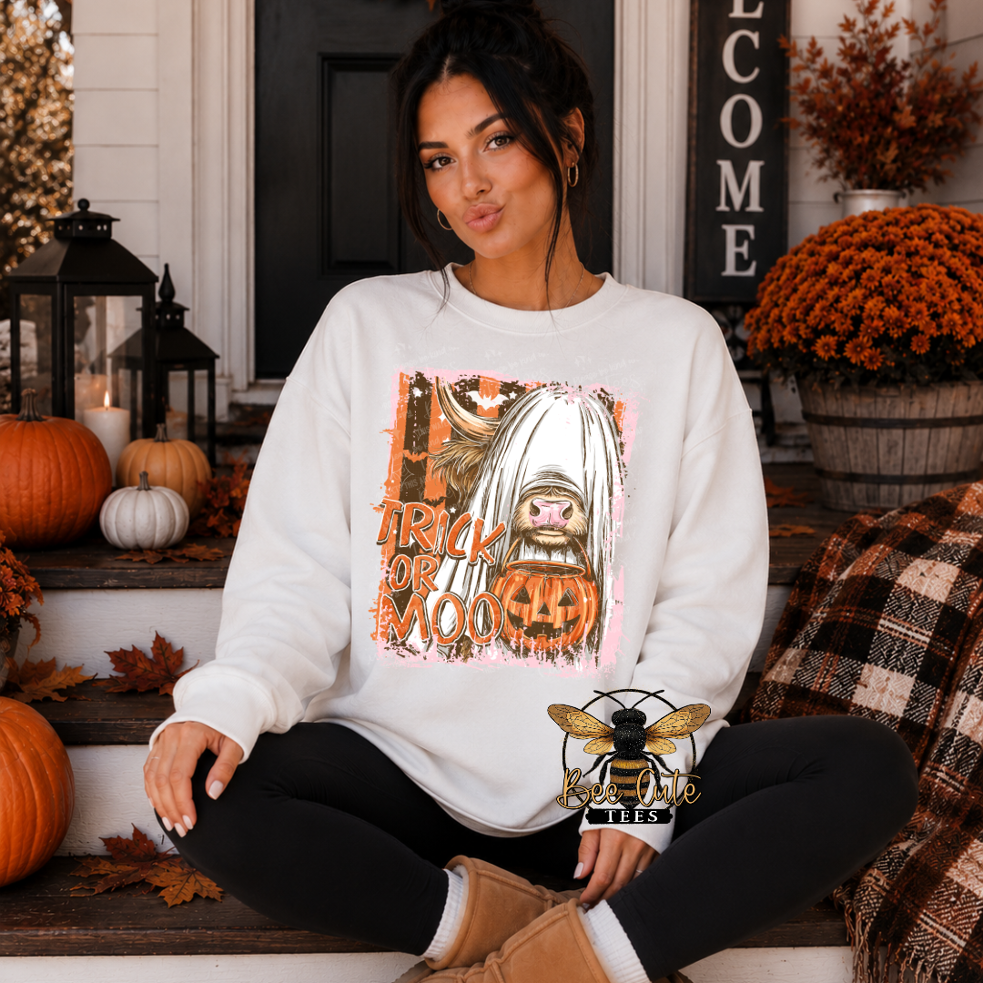A woman sits on outdoor porch steps wearing a white crewneck sweatshirt with a Halloween-themed graphic print featuring a ghost, a jack-o'-lantern, and the text 'TRICK OR MOO'. The background is decorated with pumpkins, mums, and fall leaves.
