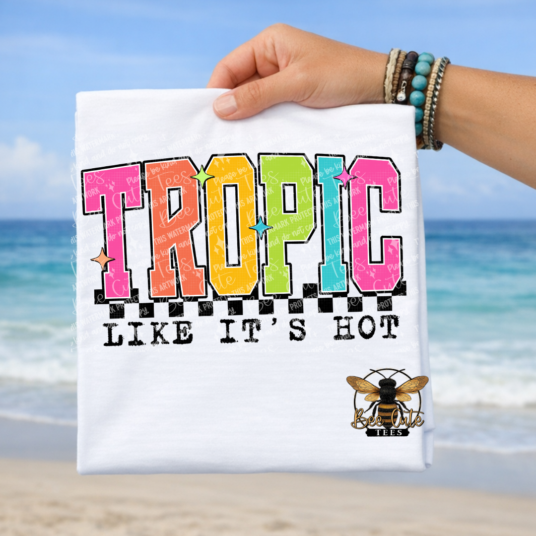 Hand holding a white t-shirt with 'Tropic Like It's Hot' text and a bee logo against a beach background.