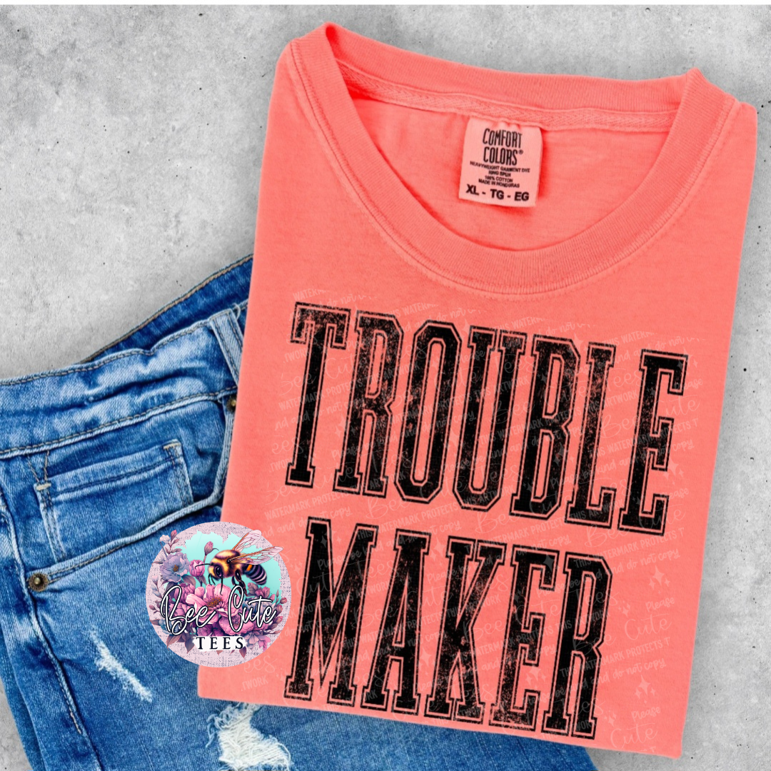 Pink t-shirt with 'Trouble Maker' text on a concrete surface with jeans and a sticker.