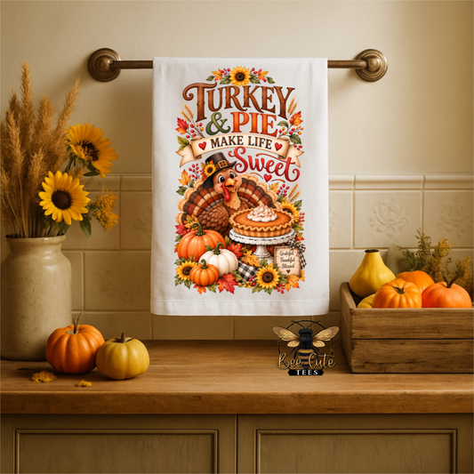 A white kitchen towel with a colorful Thanksgiving-themed graphic hanging on a brass towel bar in a kitchen setting. The graphic reads 'Turkey & Pies Make Life Sweet' with an illustration of a turkey, pie, pumpkins, sunflowers, and autumn leaves.